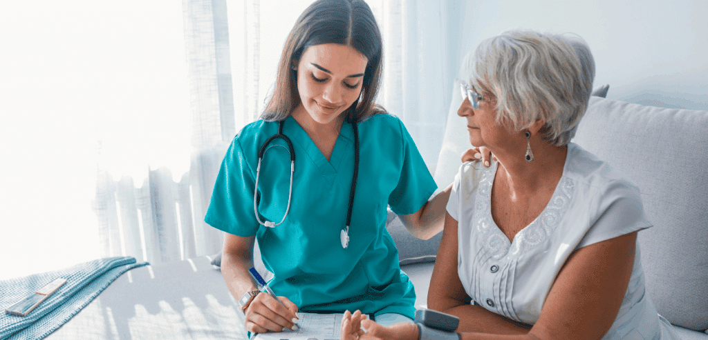 A nurse providing home health to her patient. She is sitting next to a female patient on a white bed. The nurse is writing on a clipboard and smiling while the client looks at her.