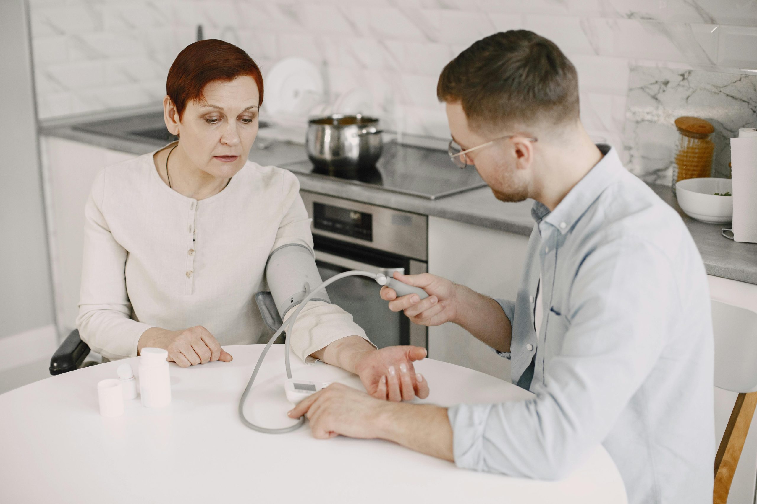 Adult man monitoring elderly woman's blood pressure at kitchen table using sphygmomanometer.