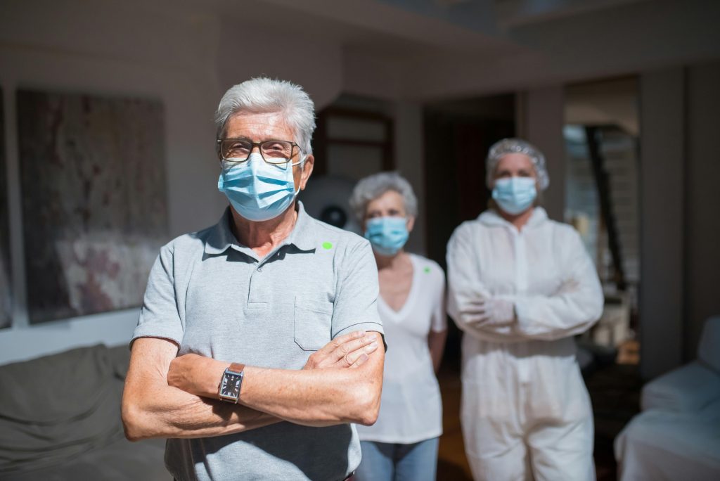 Elderly couple and healthcare professional in PPE wearing face masks indoors, reflecting health precautions.