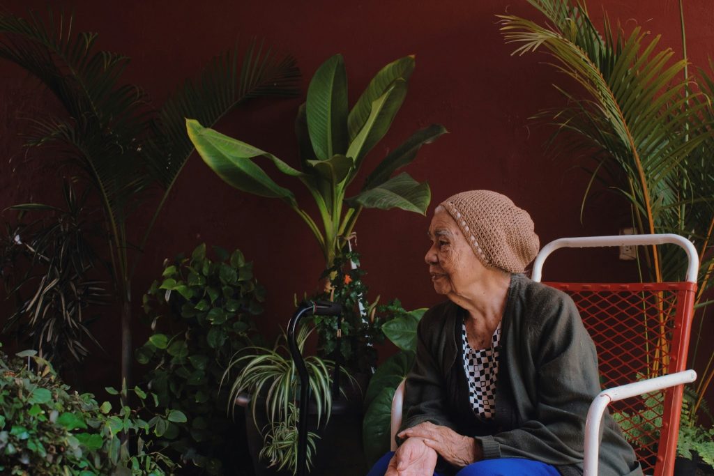 Elderly woman sitting in red chair with a background of plants.